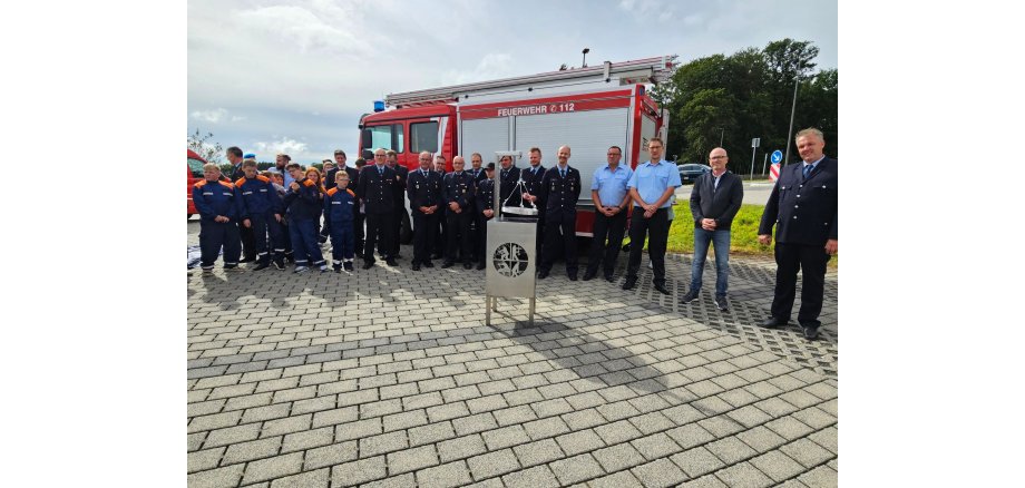 Eine Gruppe von Menschen steht vor einem Feuerwehrfahrzeug im Halbkreis. Im Vordergrund ein Grill
