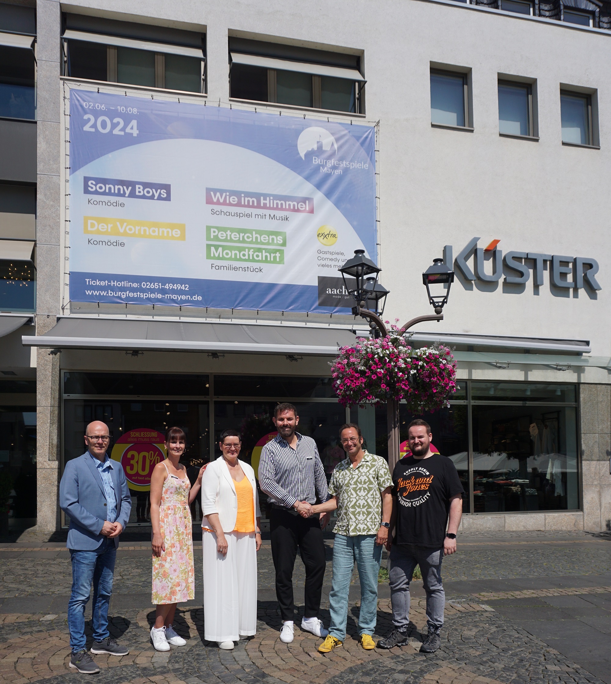 v. l. n. r.: Oberbürgermeister Dirk Meid, Leiterin der Jungen Mode Katrin Fuhrmann, Leiterin DOB Elke Münch, Leiter der Herrenabteilung Ralf Sturtz, Intendant Alexander May und Verwaltungsleiter der Burgfestspiele Kevin Stoye vor dem Plakat am Aachener Modehaus
