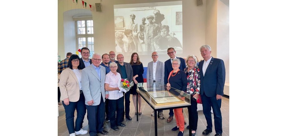 Mehrere Personen stehen um einen Glaskasten - im Hintergrund ein Bild in schwarz-weiß