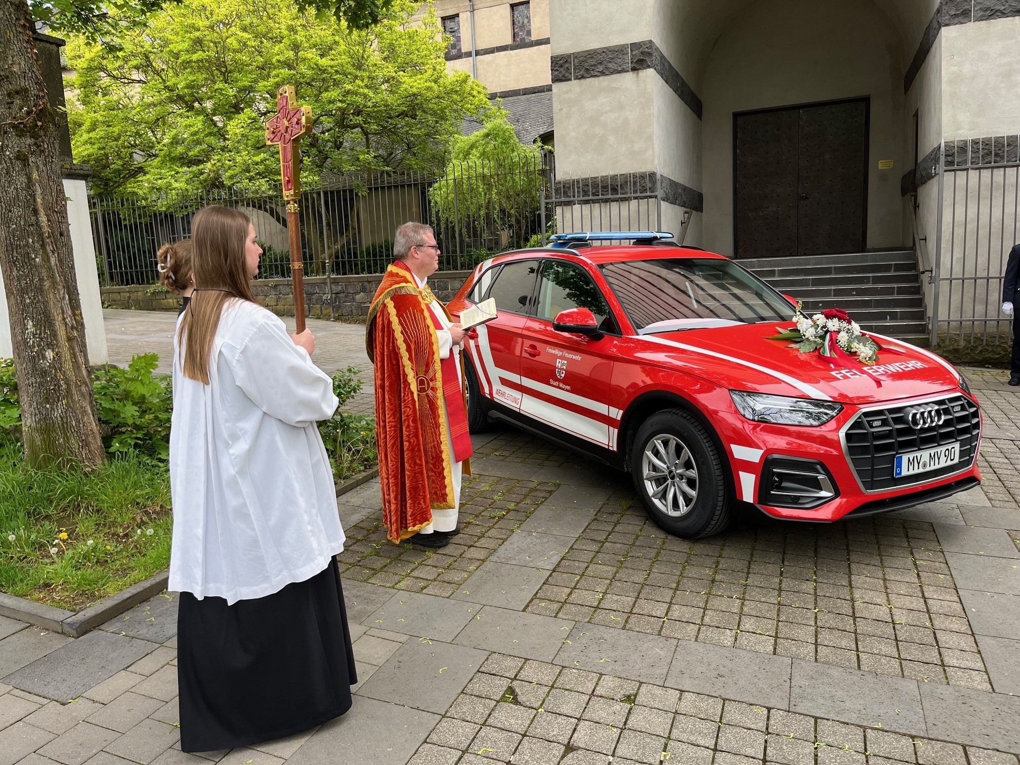 Maiübung der Freiwilligen Feuerwehr Mayen und Einsegnung des Kommandowagens Pastor segnet einen Wagen ein, da neben steht eine Messdienerin