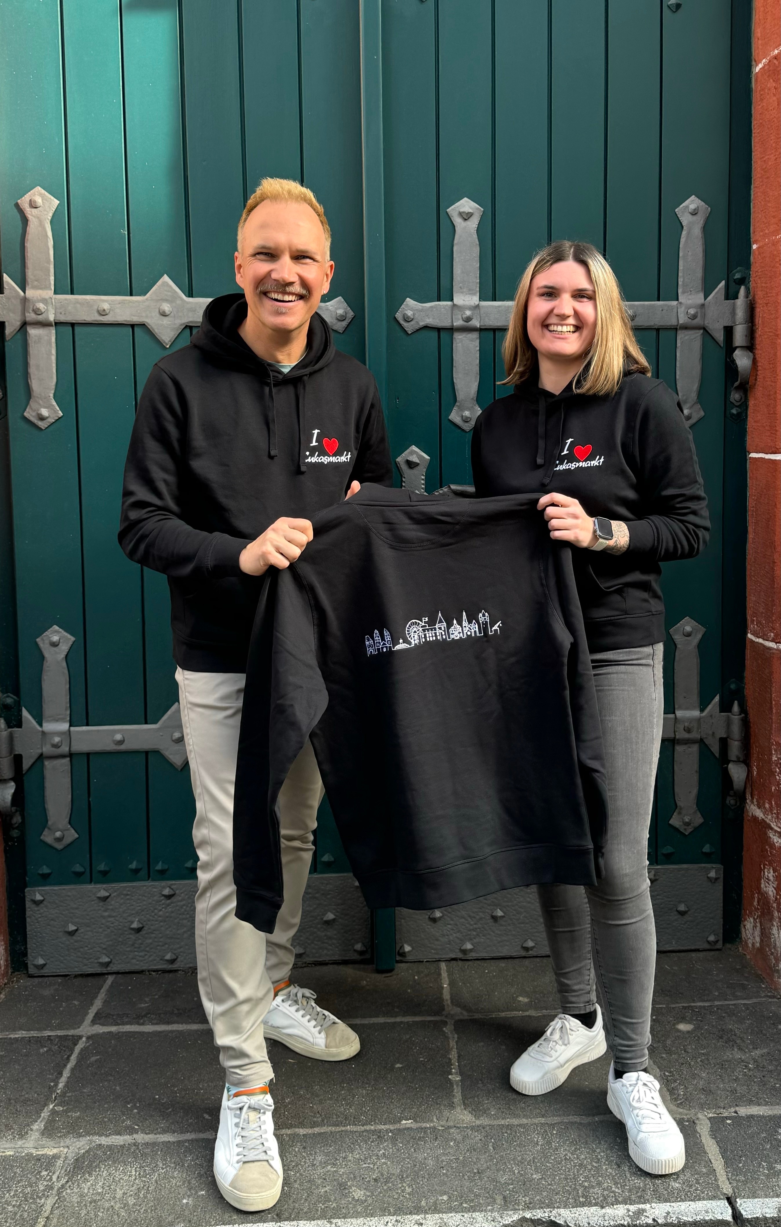 Eine Frau und ein Mann halten zusammen einen Pullover vor sich und präsentieren diesen