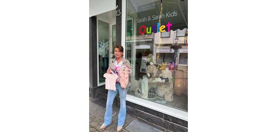 Frau steht mit 2 Kleidungsstücken für Babys in der Hand vor einem Laden