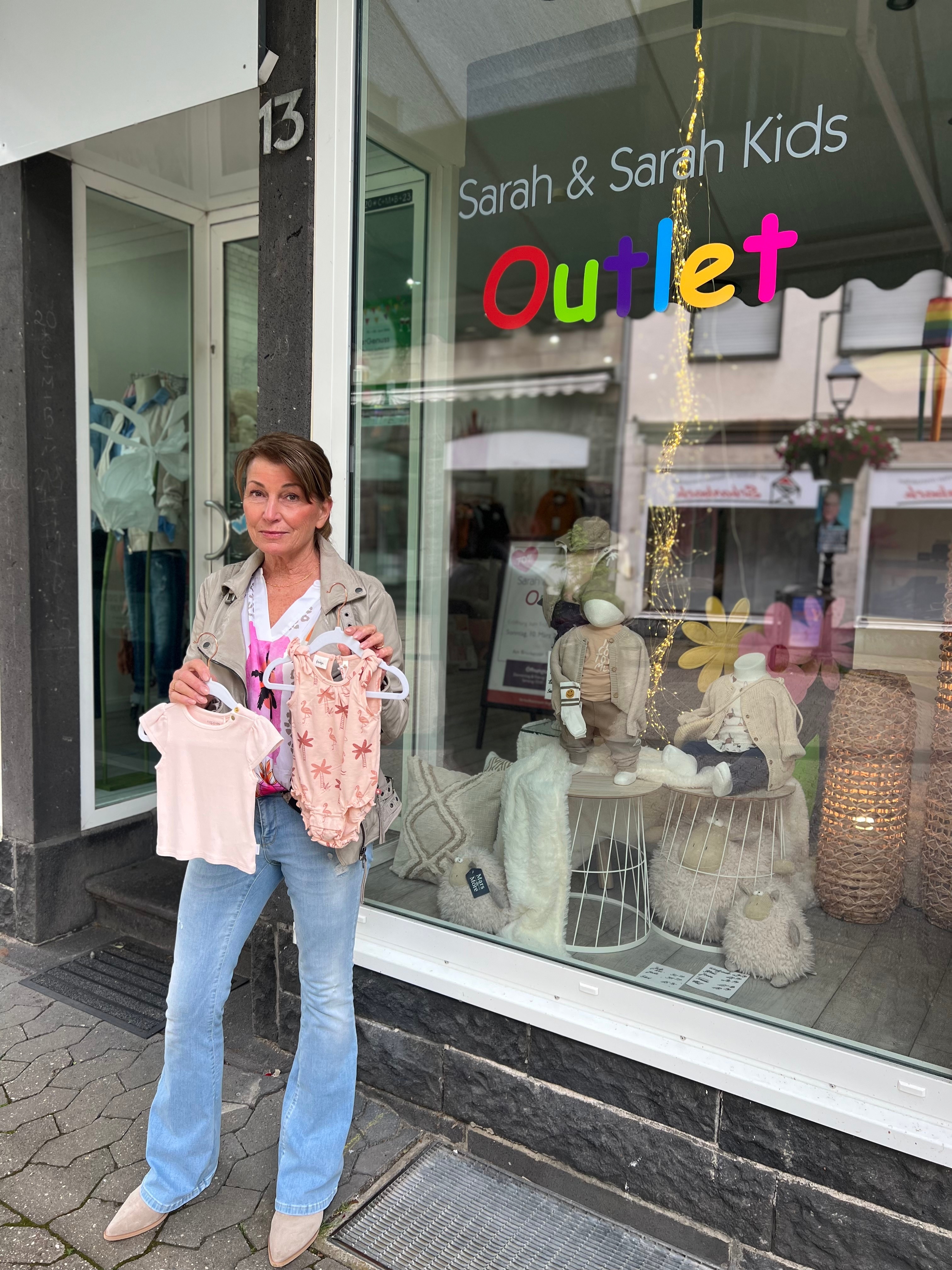 Frau steht mit 2 Kleidungsstücken für Babys in der Hand vor einem Laden