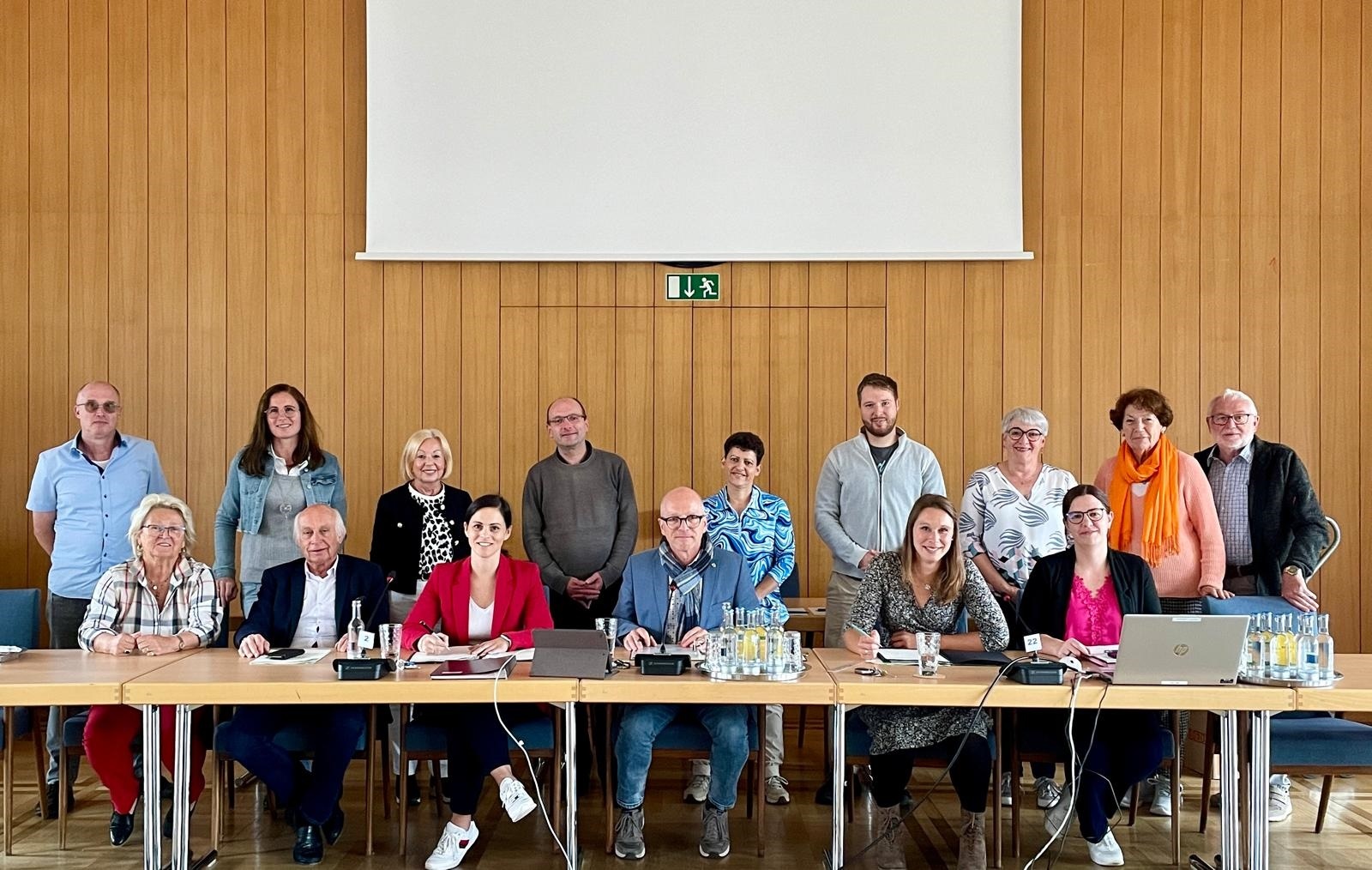 Mehrere Personen, einige sitzend, einige stehend im Sitzungssaal vor einer Leinwand