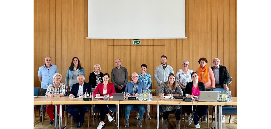 Mehrere Personen, einige sitzend, einige stehend im Sitzungssaal vor einer Leinwand