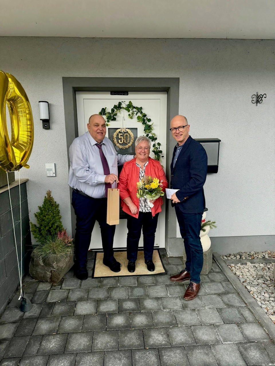 2 Männer und eine Frau stehe vor einer Haustür und haben Präsente in den Händen