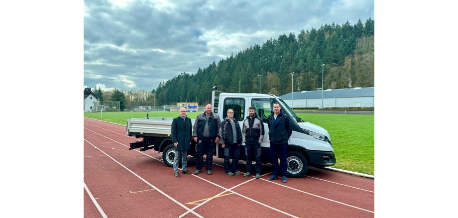 5 Herren stehen vor einem Wagen auf der Laufbahn eines Sportplatzes