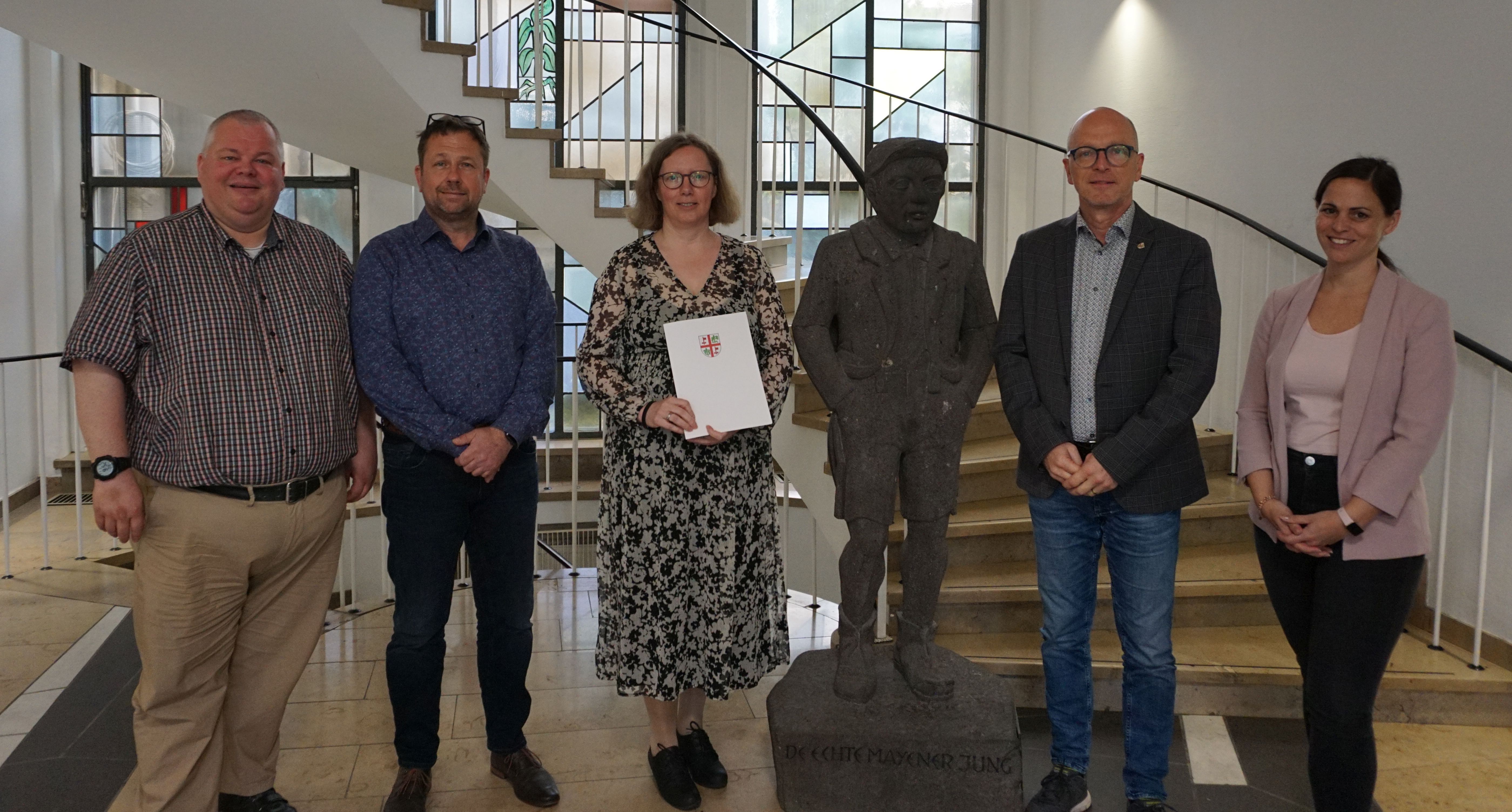 5 Personen stehen im Treppenhaus - dazwischen eine Steinfigur. Die Dame in der Mitte hält eine Urkunde in den Händen