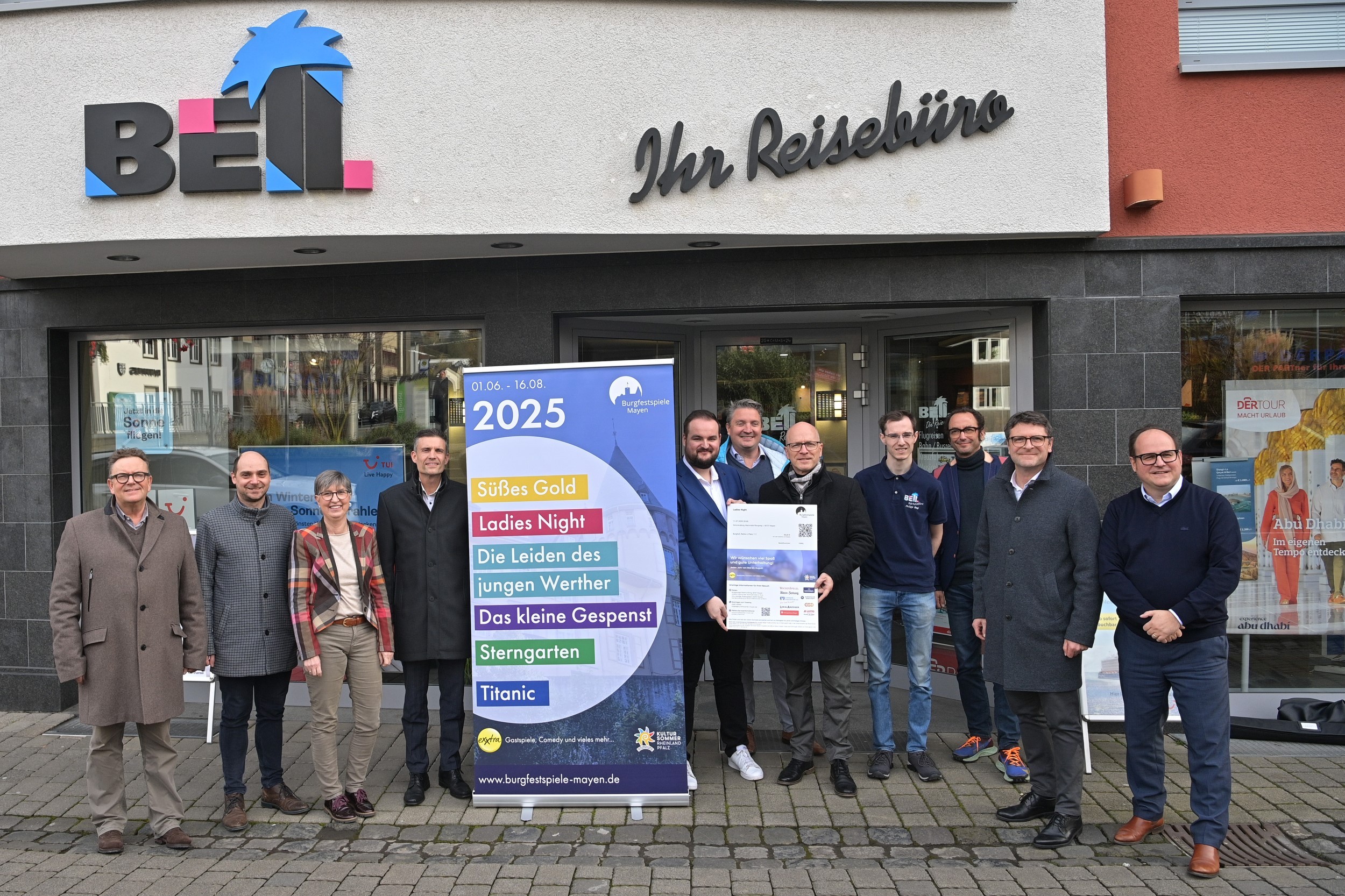 11 Personen vor dem Reisebüro Bell. Zwischen ihnen steht ein Plakat mit den Theaterstücken der Burgfestspiele 2025