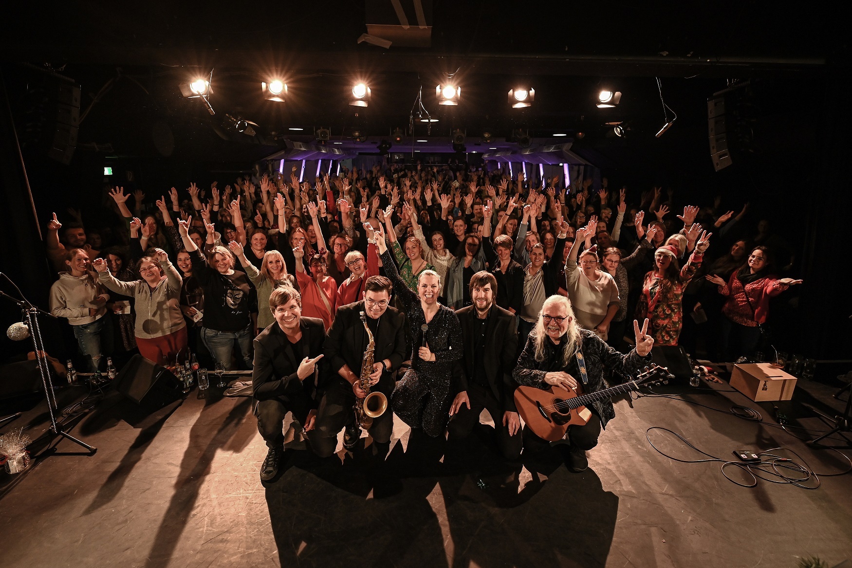 5 Personen stehen mit Instrumenten auf der Bühne - im Hintergrund viele Menschen, die die Arme hoch werfen