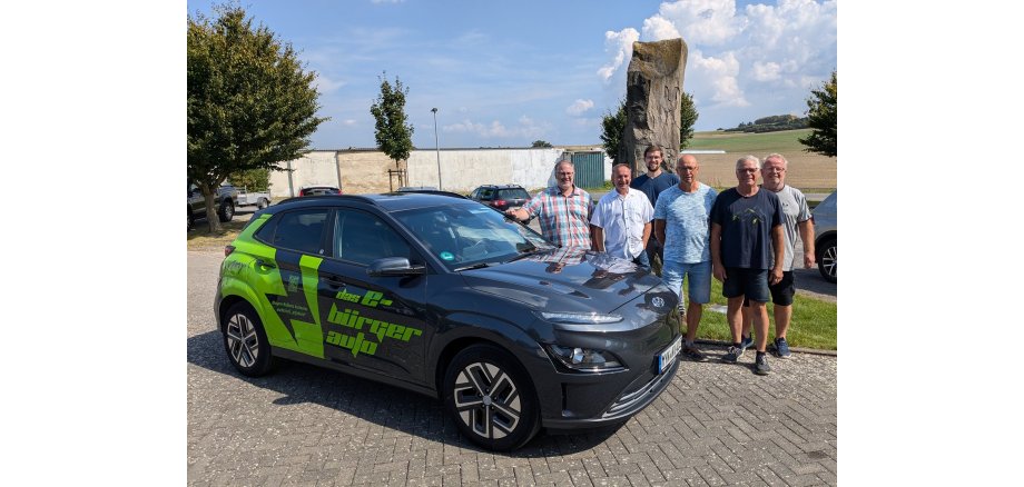 Ortsvorsteher Sven Weber (ganz links), Klimaschutzmanager Helge Lippert (3. Von links) zusammen mit den vier Kümmerern präsentieren das Dorfauto am Bürgerhaus Hausen 6 Männer stehen neben einem E-Auto