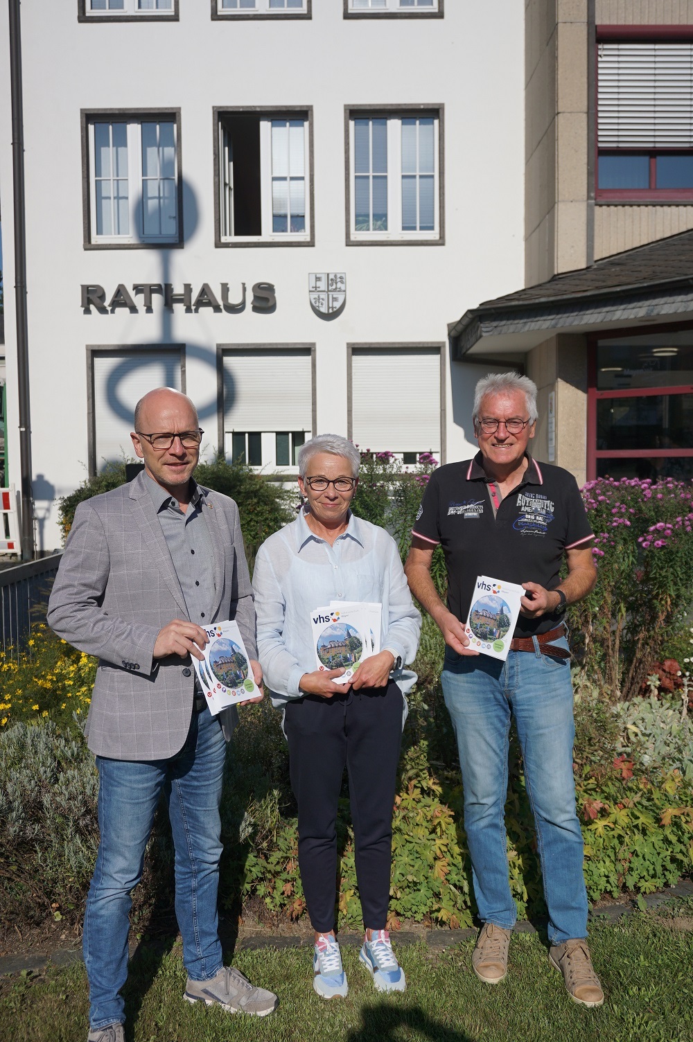 Zwei Männer und eine Frau stehen vor dem Rathaus und halten VHS-Hefte in den Händen