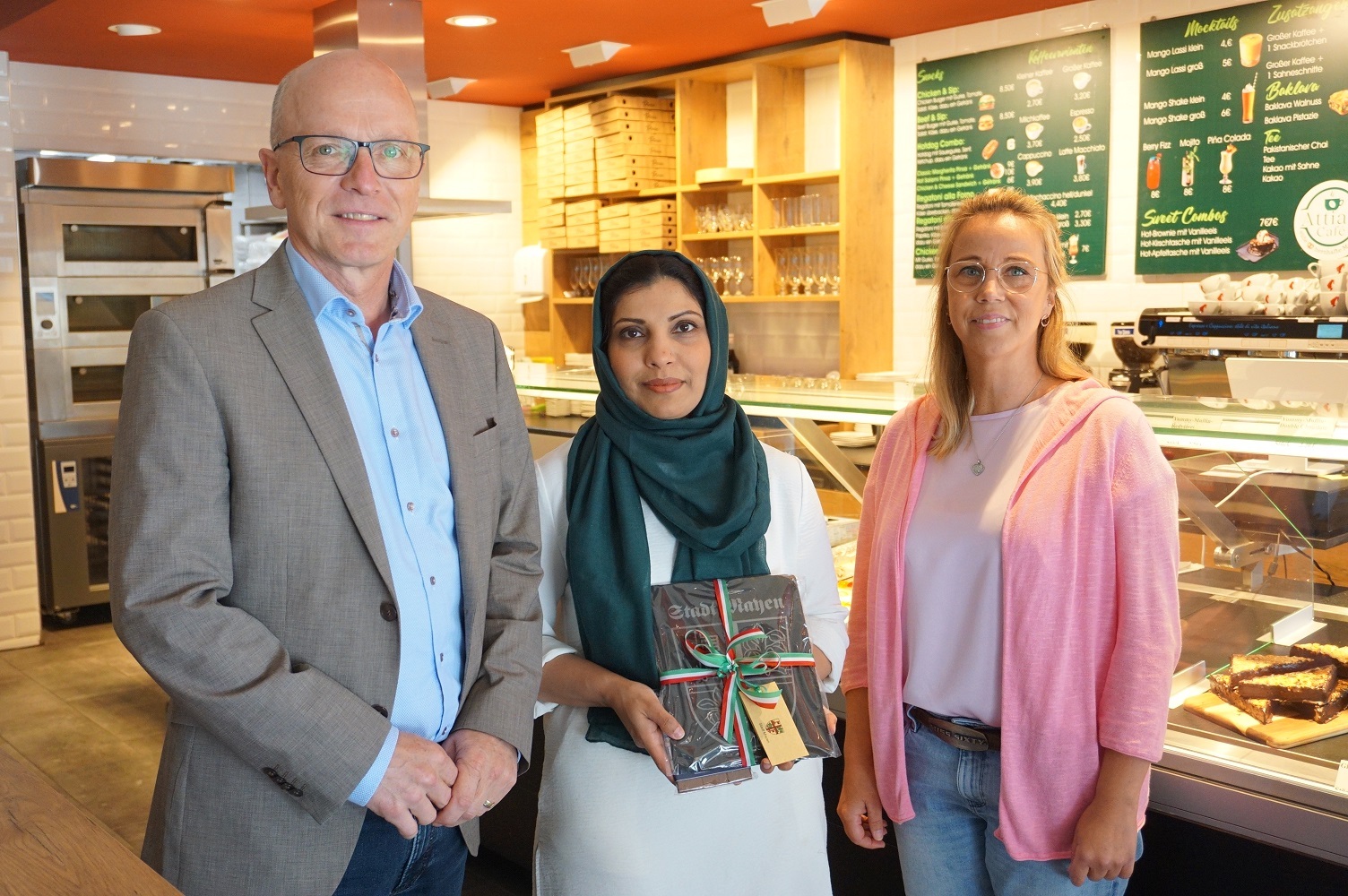 3 Personen stehen vor einer Theke - die Dame in der Mitte hält ein Präsent in den Händen