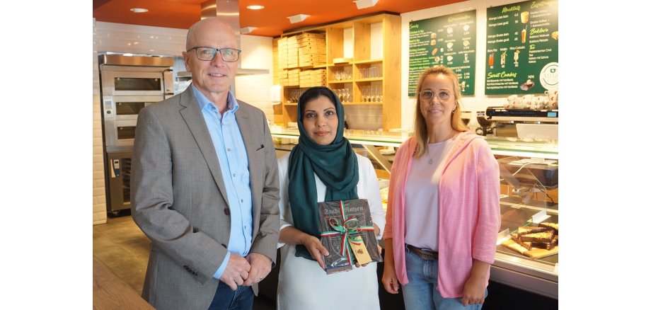 3 Personen stehen vor einer Theke - die Dame in der Mitte hält ein Präsent in den Händen