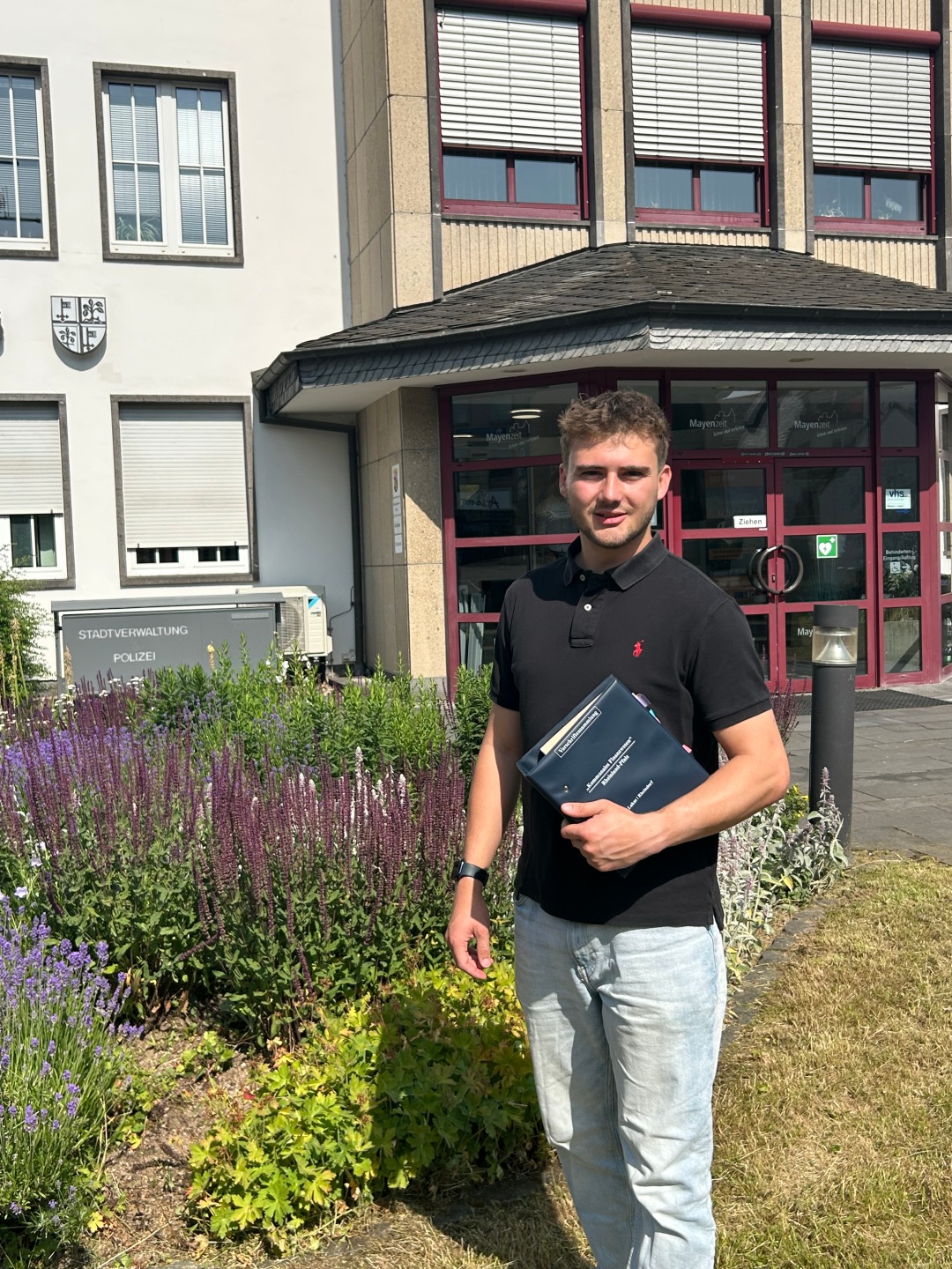 Marius Luxem befindet sich aktuell im dualen Studium zum Stadtinspektor. Marius Luxem hält ein Gesetzbuch in der Hand und steht vor der Stadtverwaltung