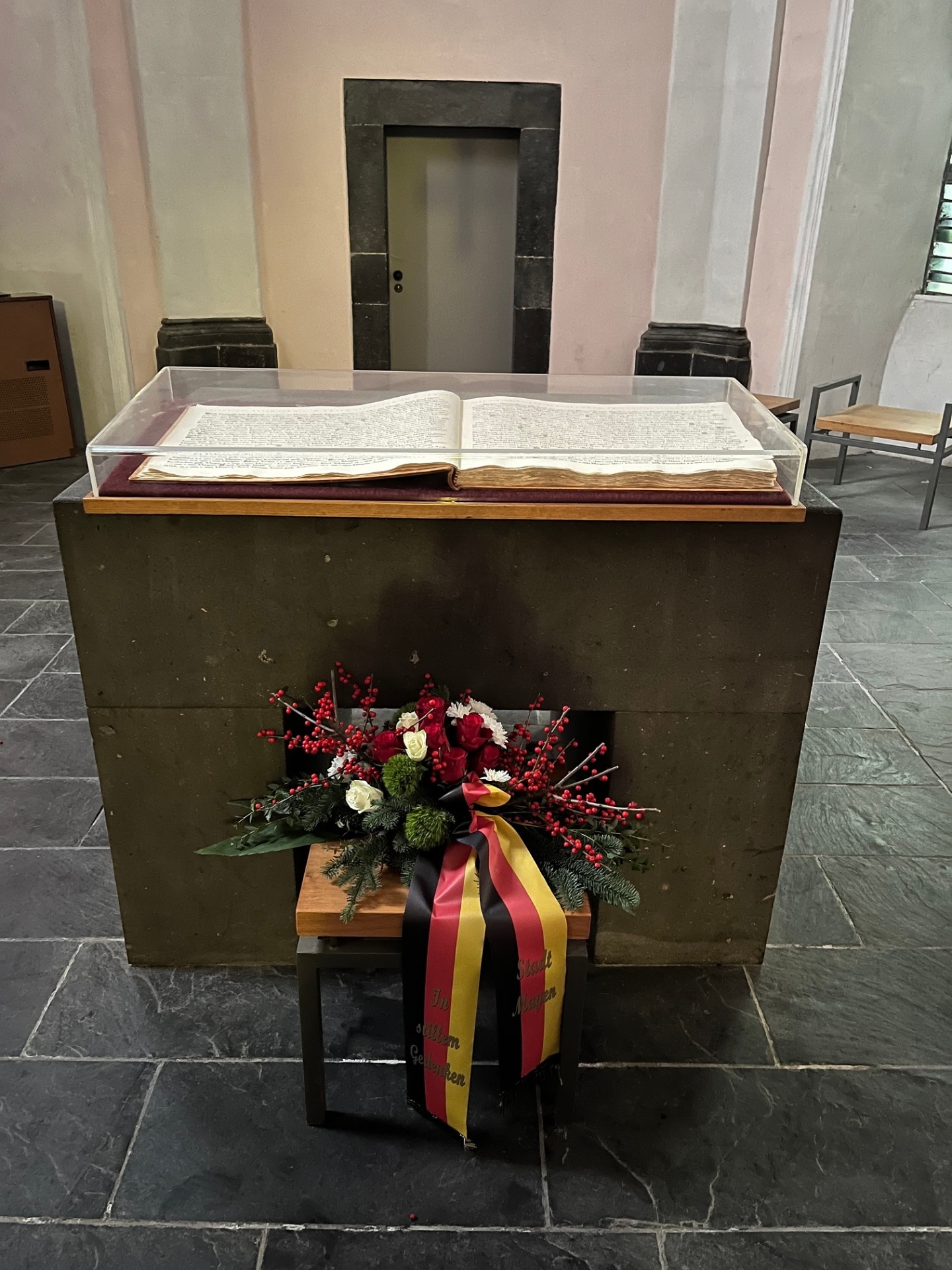 Aufgeschlagenes Buch auf einem Sockel. Davor steht ein Blumengesteck