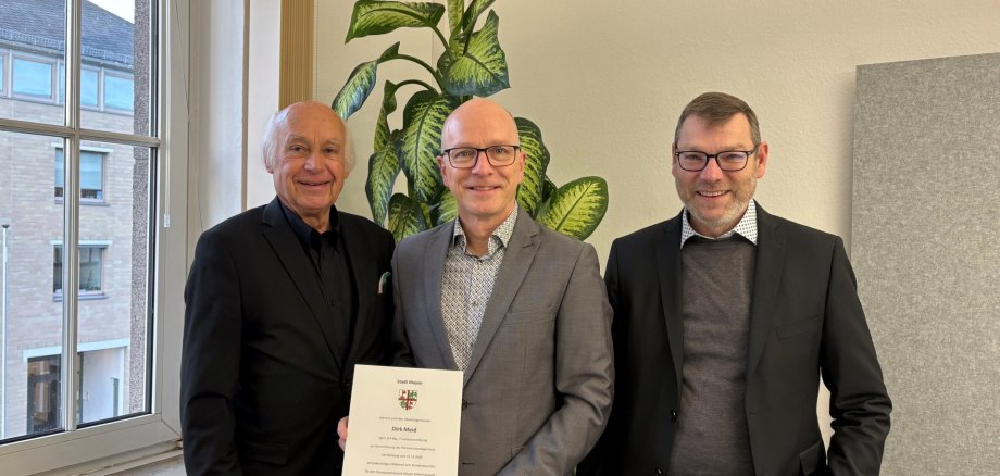 Bürgermeister Bernhard Mauel, Oberbürgermeister Dirk Meid und Beigeordneter Thomas Schroeder stehen neben einem Fenster vor einer Pflanze. OB Meid hält seine Urkunde in der Hand