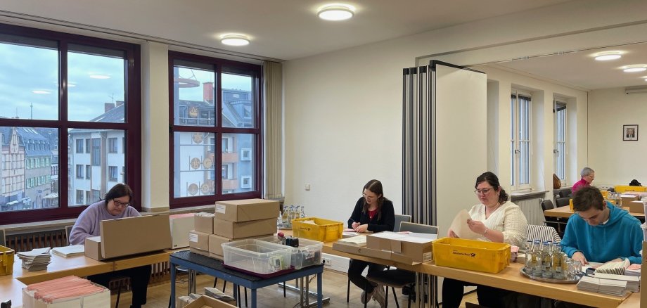 Zahlreiche helfende Hände bereiten die Briefwahlunterlagen für den Versand vor Mehrere Personen sitzen an Tischen und bereiten die Briefwahlunterlagen vor