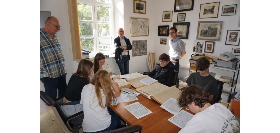 Oberbürgermeister Dirk Meid beobachtet gemeinsam mit Lehrer Edwin Kleis und Archivar Stefan Wilden die Schülerinnen und Schüler bei der Quellenarbeit Mehrere Personen sitzen und stehen an einem Tisch und recherchieren
