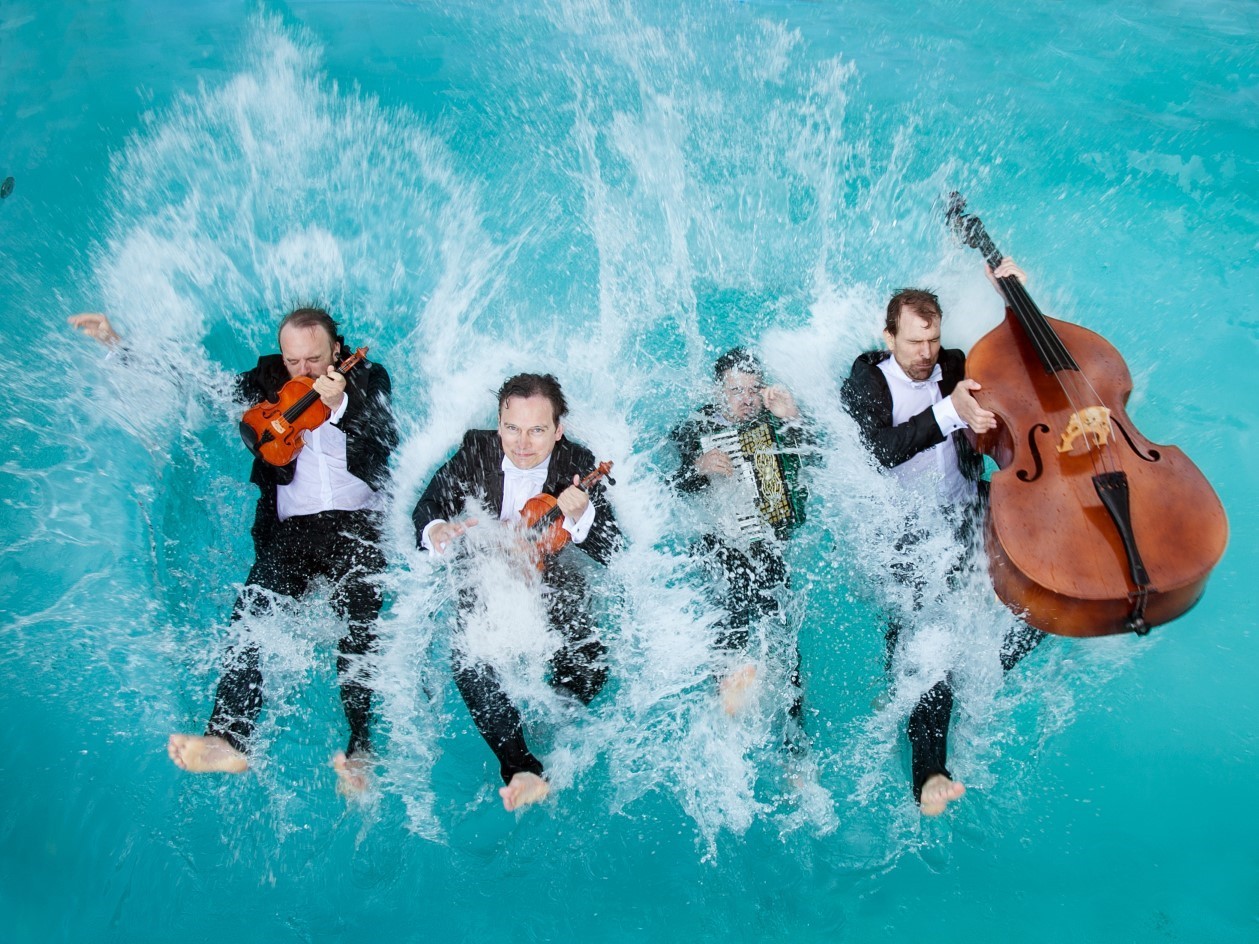Vier Männer in Anzug und mit Instrumenten fallen rückwärts ins Wasser
