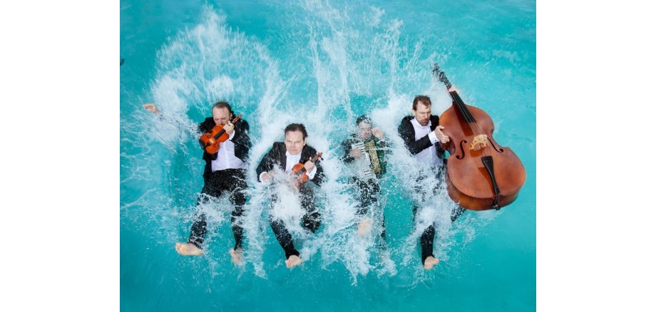 Vier Männer in Anzug und mit Instrumenten fallen rückwärts ins Wasser