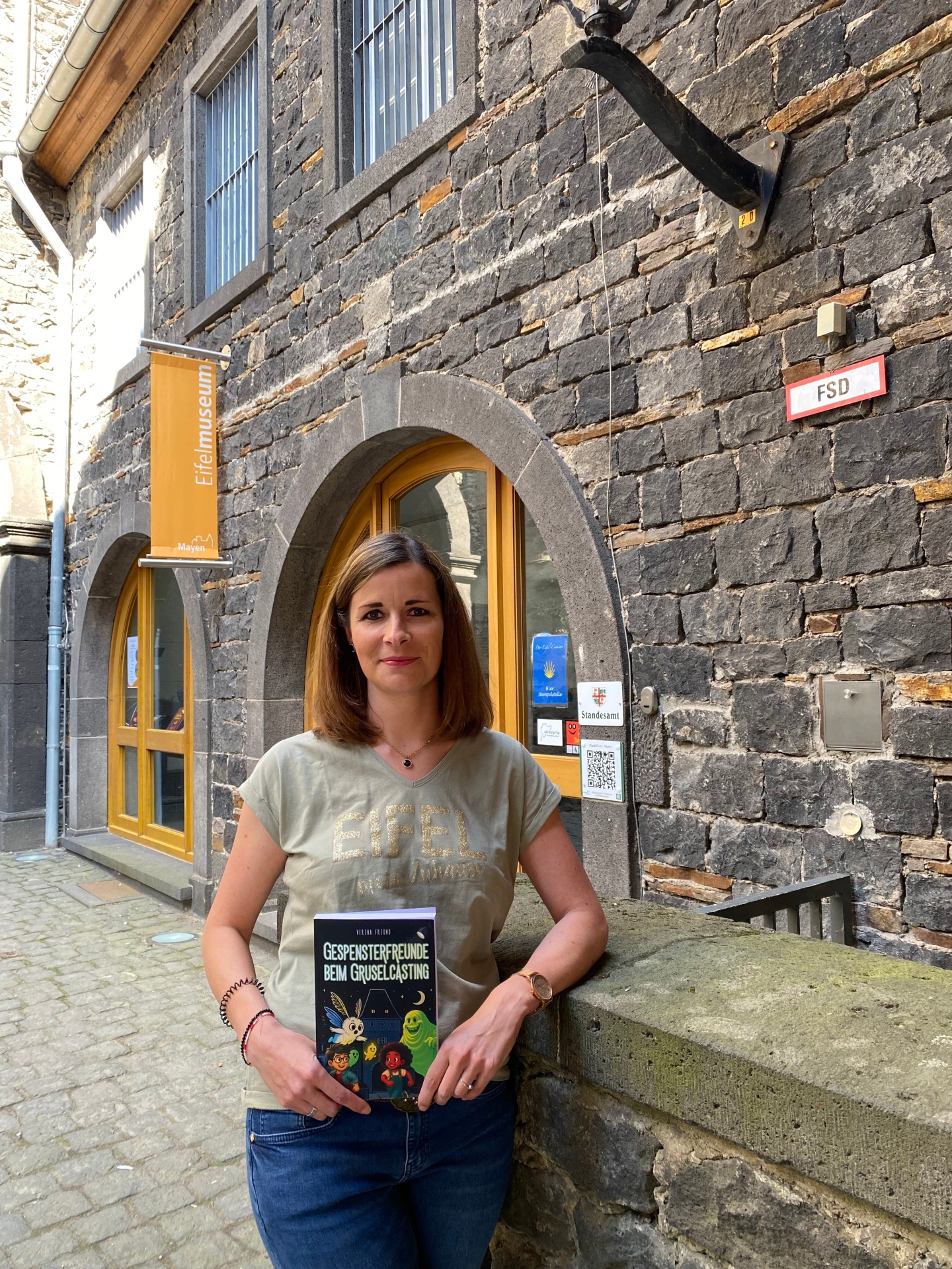 Kinderbuchautorin Verena Freund-Donnhauser steht mit ihrem Buch vor dem Eingang des Eifelmuseums
