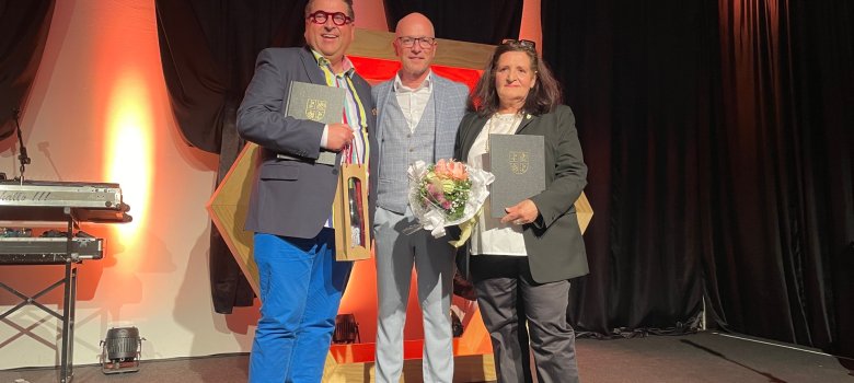 Oberbürgermeister Dirk Meid überreichte Judith Ellner und Frank Gondorf die Goldene Ehrennadel der Stadt Mayen Drei Personen mit Präsenten auf der Bühne