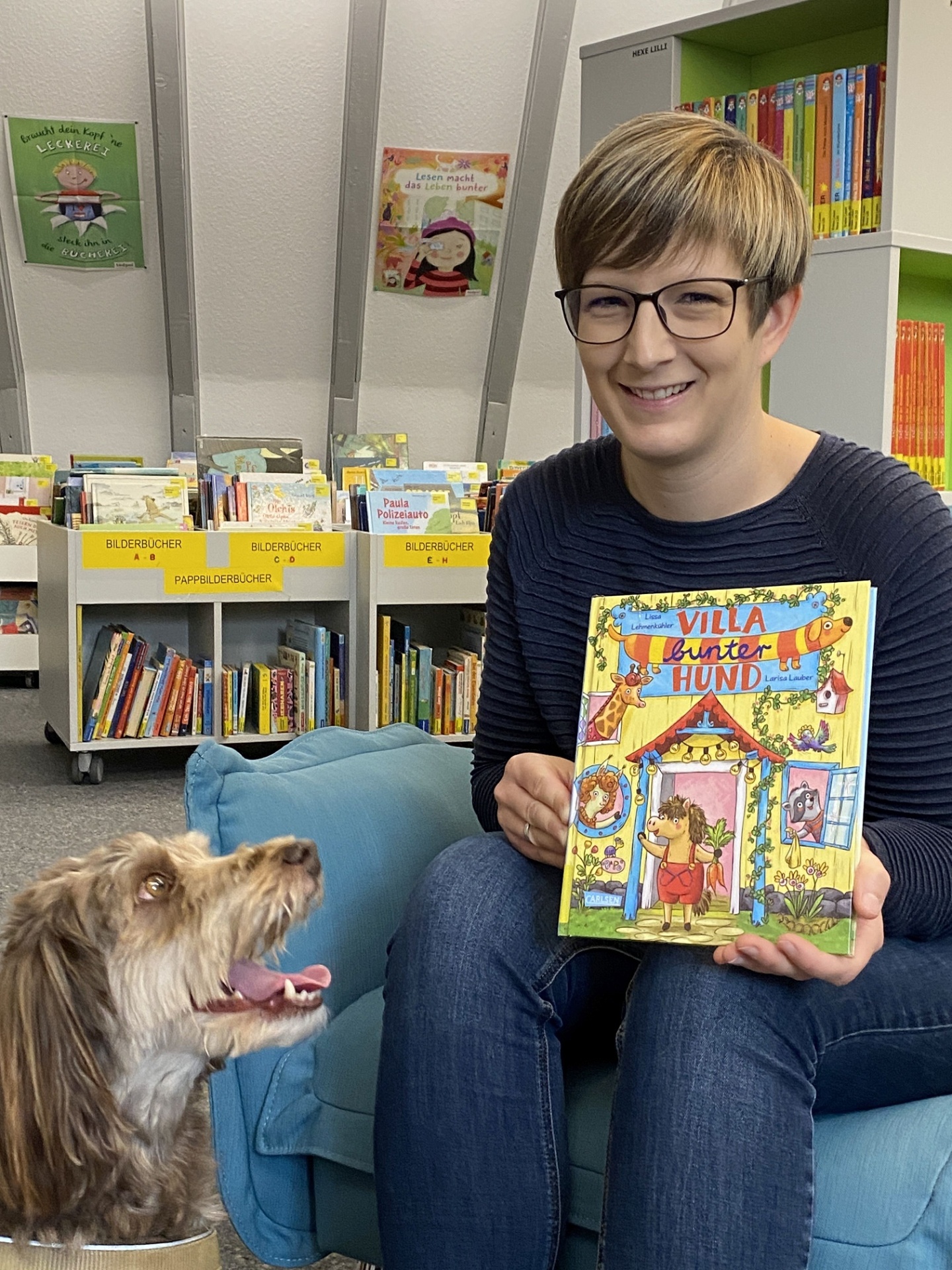 Svenja Weinand sitzt mit einem bunten Kinderbuch in der Hand auf einem Sessel in der Kinderbücherei. Neben ihr sitzt ein brauner Hund und schaut sie an.