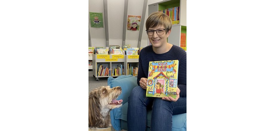 Svenja Weinand sitzt mit einem bunten Kinderbuch in der Hand auf einem Sessel in der Kinderbücherei. Neben ihr sitzt ein brauner Hund und schaut sie an.