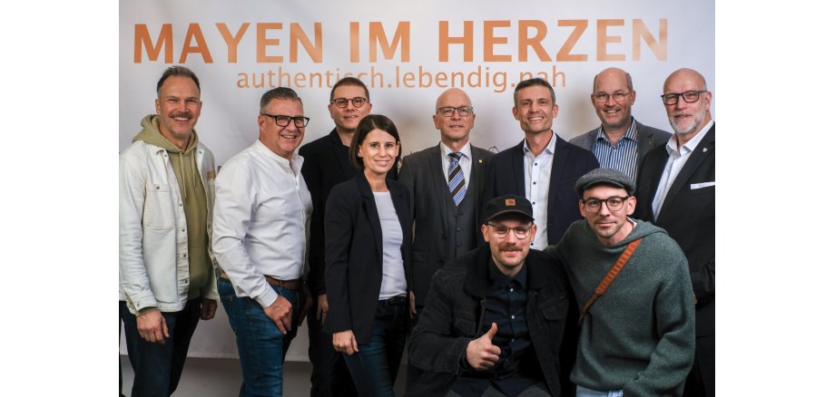 Kinopremiere feiert neue Imagekampagne Neun Männer und eine Frau stehen vor einer Wand mit orangener "Mayen im Herzen" Aufschrift