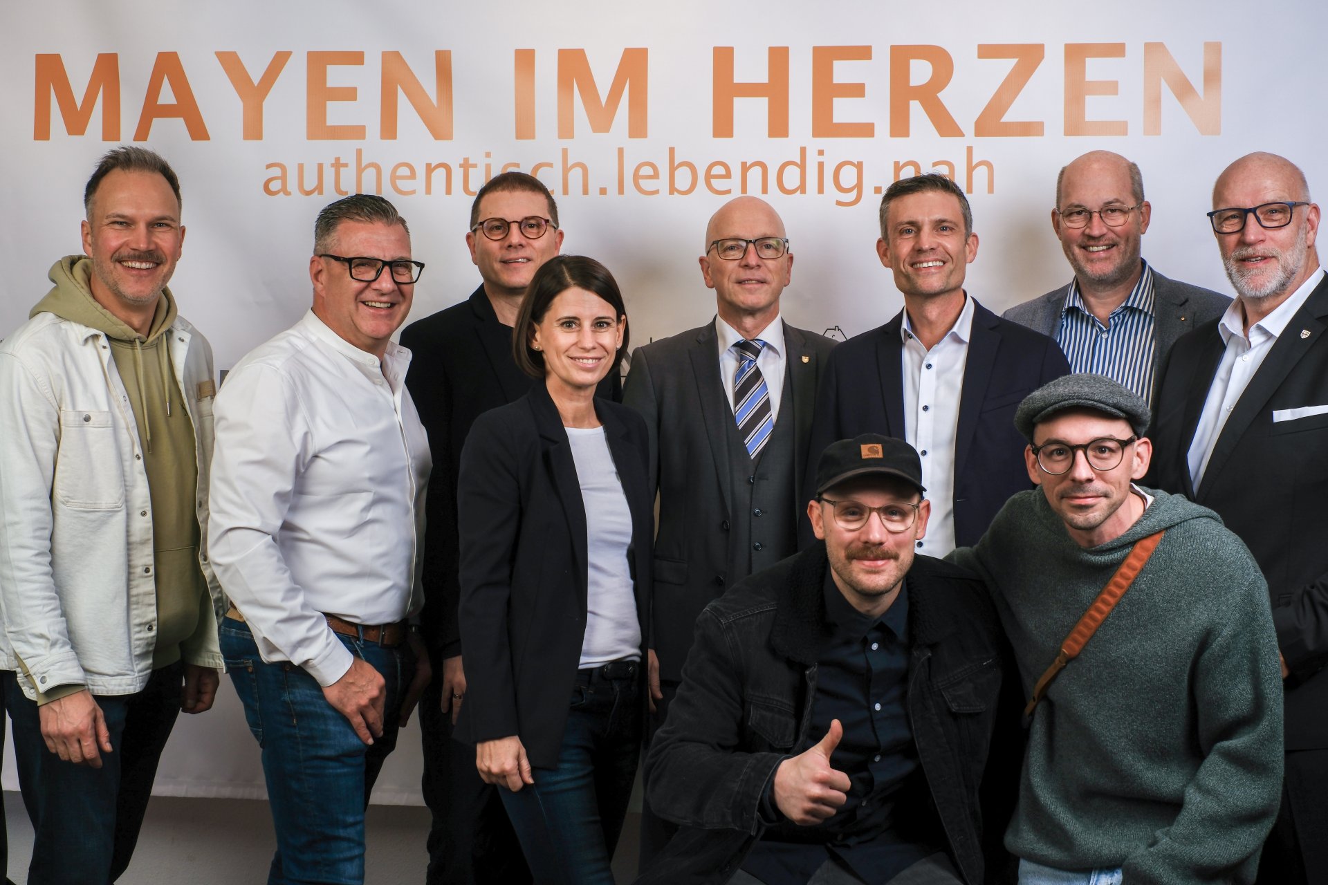Neun Männer und eine Frau stehen vor einer Wand mit orangener "Mayen im Herzen" Aufschrift