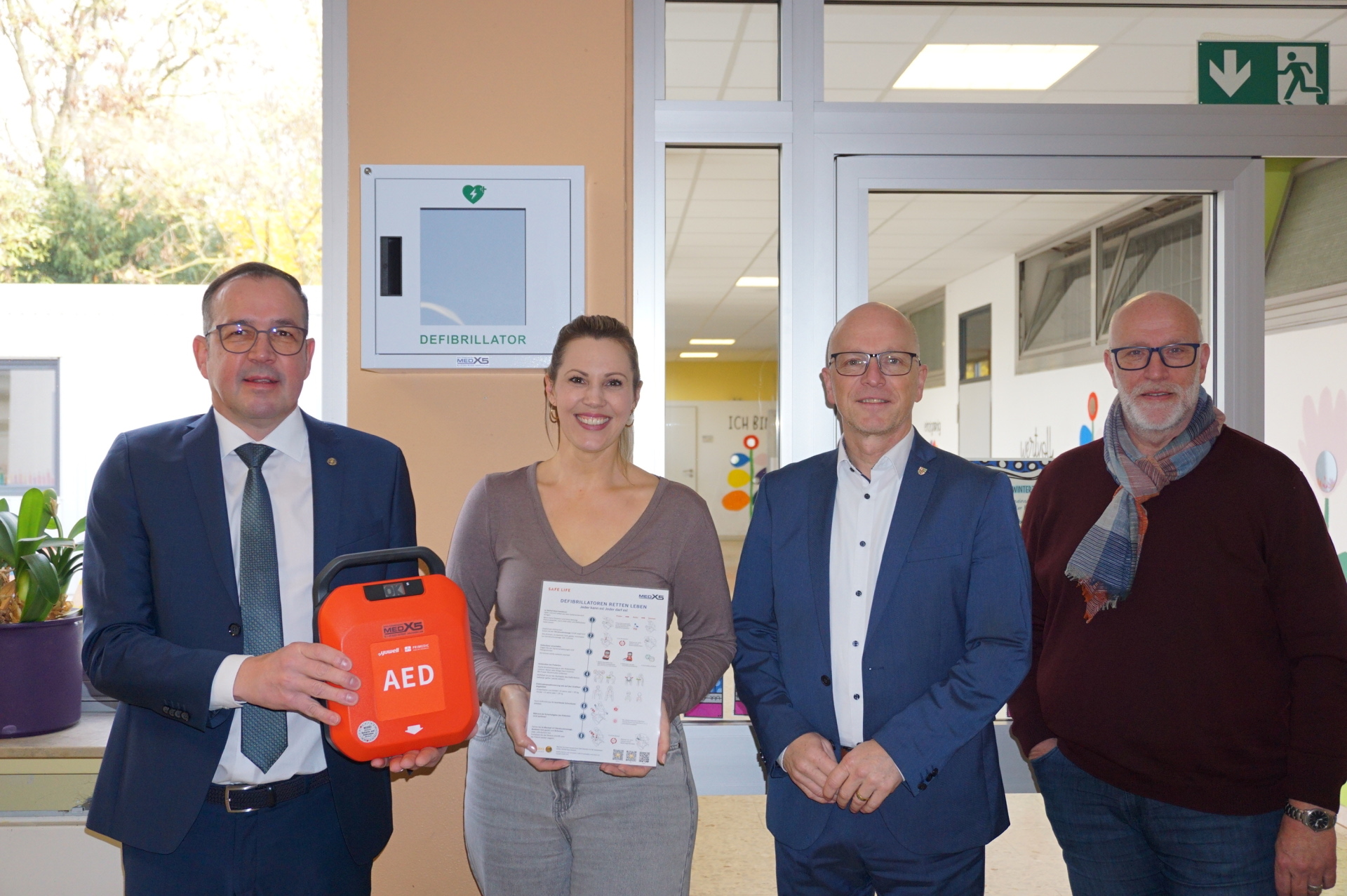 Markus Schmitt vom Rotary Club, Direktorin Ann-Marlen Schreiber, Oberbürgermeister Dirk Meid und Fachbereichsleiter Uwe Hoffmann stehen gemeinsam vor einer Glastür. Herr Schmitt hält einen orangenen Defibrillator in der Hand