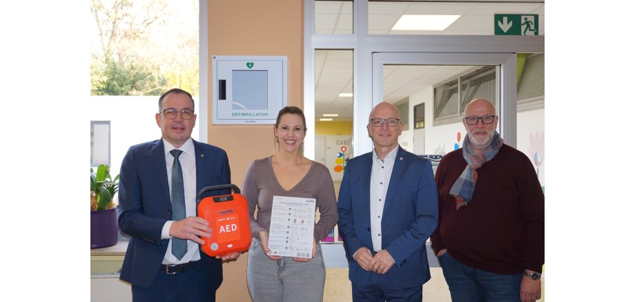 v.l.n.r.: Markus Schmitt vom Rotary Club, Direktorin Ann-Marlen Schreiber, Oberbürgermeister Dirk Meid und Fachbereichsleiter Uwe Hoffmann Markus Schmitt vom Rotary Club, Direktorin Ann-Marlen Schreiber, Oberbürgermeister Dirk Meid und Fachbereichsleiter Uwe Hoffmann stehen gemeinsam vor einer Glastür. Herr Schmitt hält einen orangenen Defibrillator in der Hand