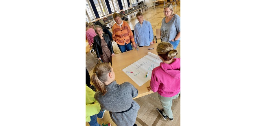 Die Mitarbeiterinnen der Stadtverwaltung Mayen bringen viele neue Ideen und Anregungen mit ein  Mehrere Personen stehen an einem Tisch zusammen - vor Ihnen ein Plakat