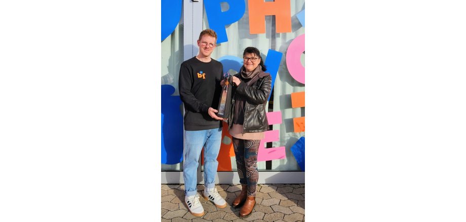 Dagmar Luxem und Benedikt Oppenhäuser stehen gemeinsam vor der bunten Eingangstür der Praxis. Dagmar Luxem Überreicht ein Präsent.