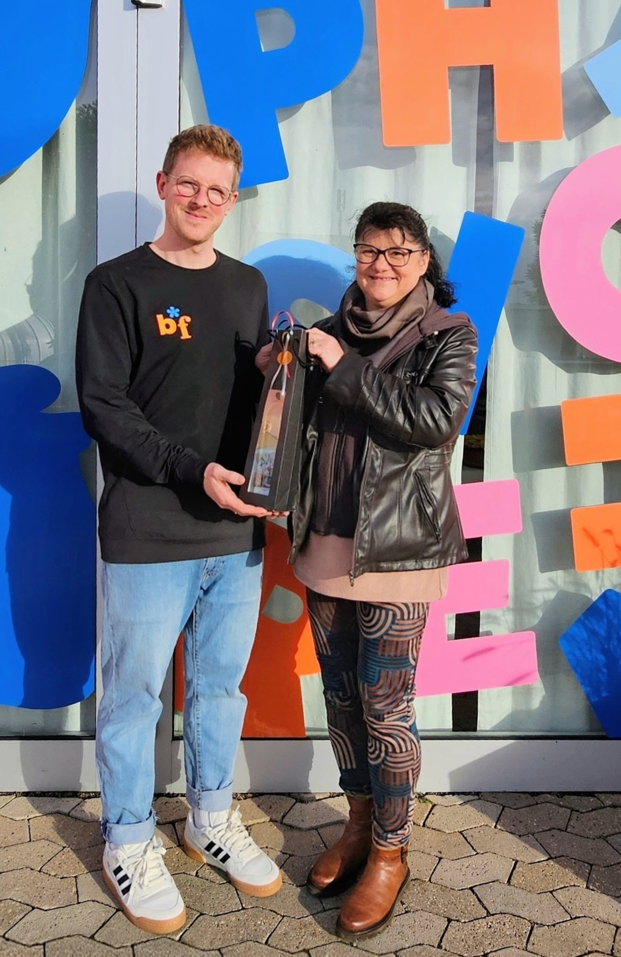 Dagmar Luxem und Benedikt Oppenhäuser stehen gemeinsam vor der bunten Eingangstür der Praxis. Dagmar Luxem Überreicht ein Präsent.