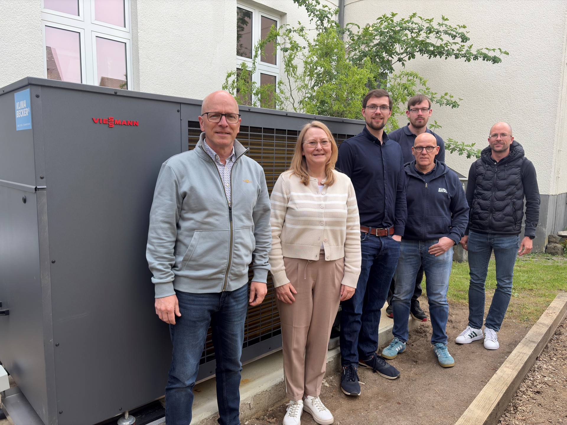 Oberbürgermeister Dirk Meid, Klimaschutzmanager Helge Lippert, Schulleiterin Simone Dumont-Knopp, Hausmeister Alexander Meurer, Fachplaner Tobias Hackenbruch, Teamleiter Klima Becker Thomas Kalischan stehen im Freien vor der Wärmepumpe