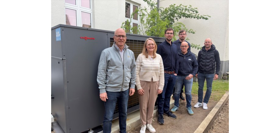 Oberbürgermeister Dirk Meid, Klimaschutzmanager Helge Lippert, Schulleiterin Simone Dumont-Knopp, Hausmeister Alexander Meurer, Fachplaner Tobias Hackenbruch, Teamleiter Klima Becker Thomas Kalischan stehen im Freien vor der Wärmepumpe