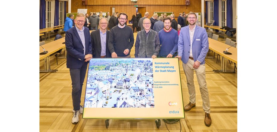 Sechs Männer stehen gemeinsam im Sitzungssaal der Stadtverwaltung hinter einem Schild mit dem Wärmeplan