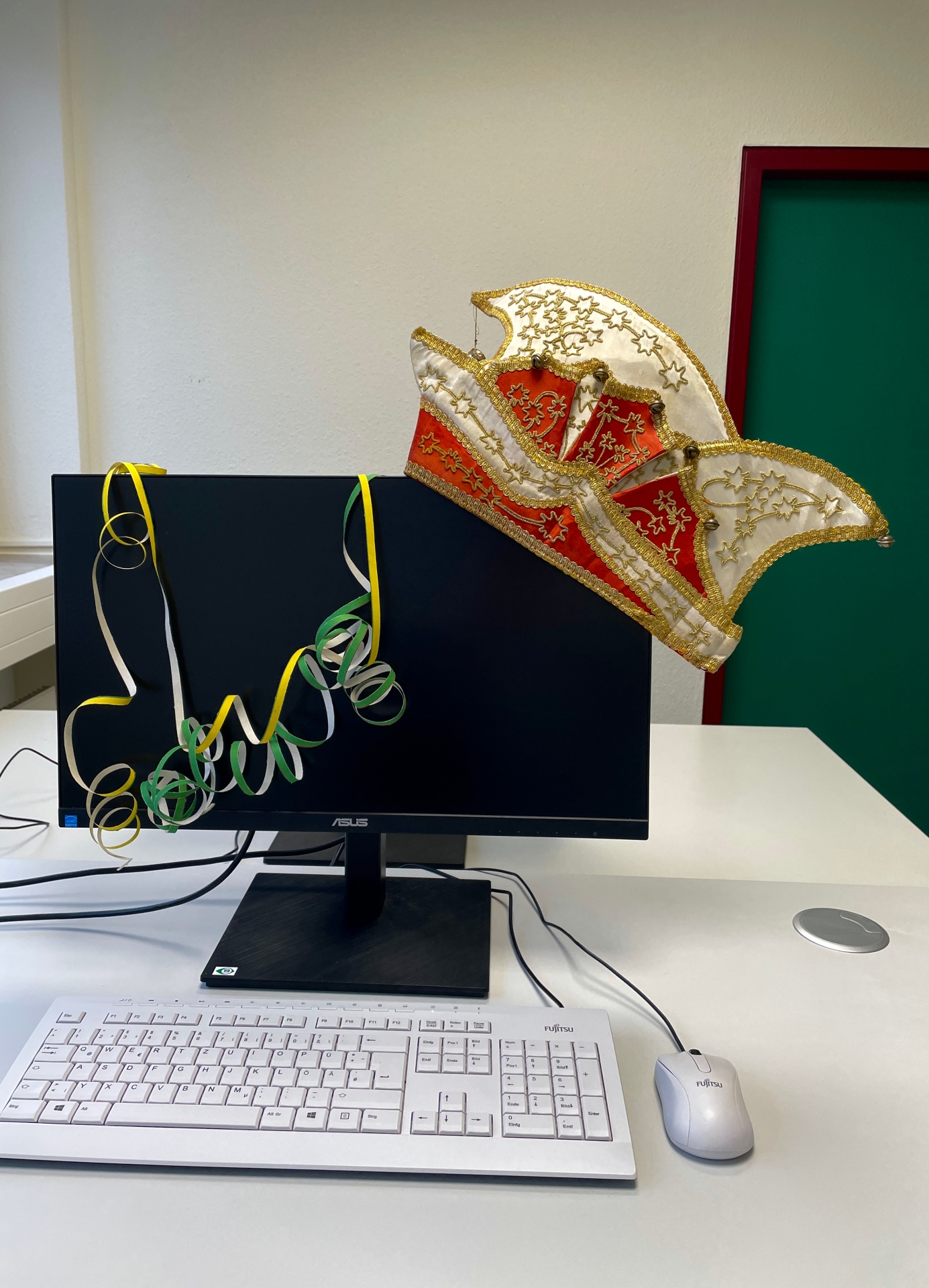 Ein bunt geschmückter Arbeitsplatz. Auf dem Monitor hängt eine Narrenmütze