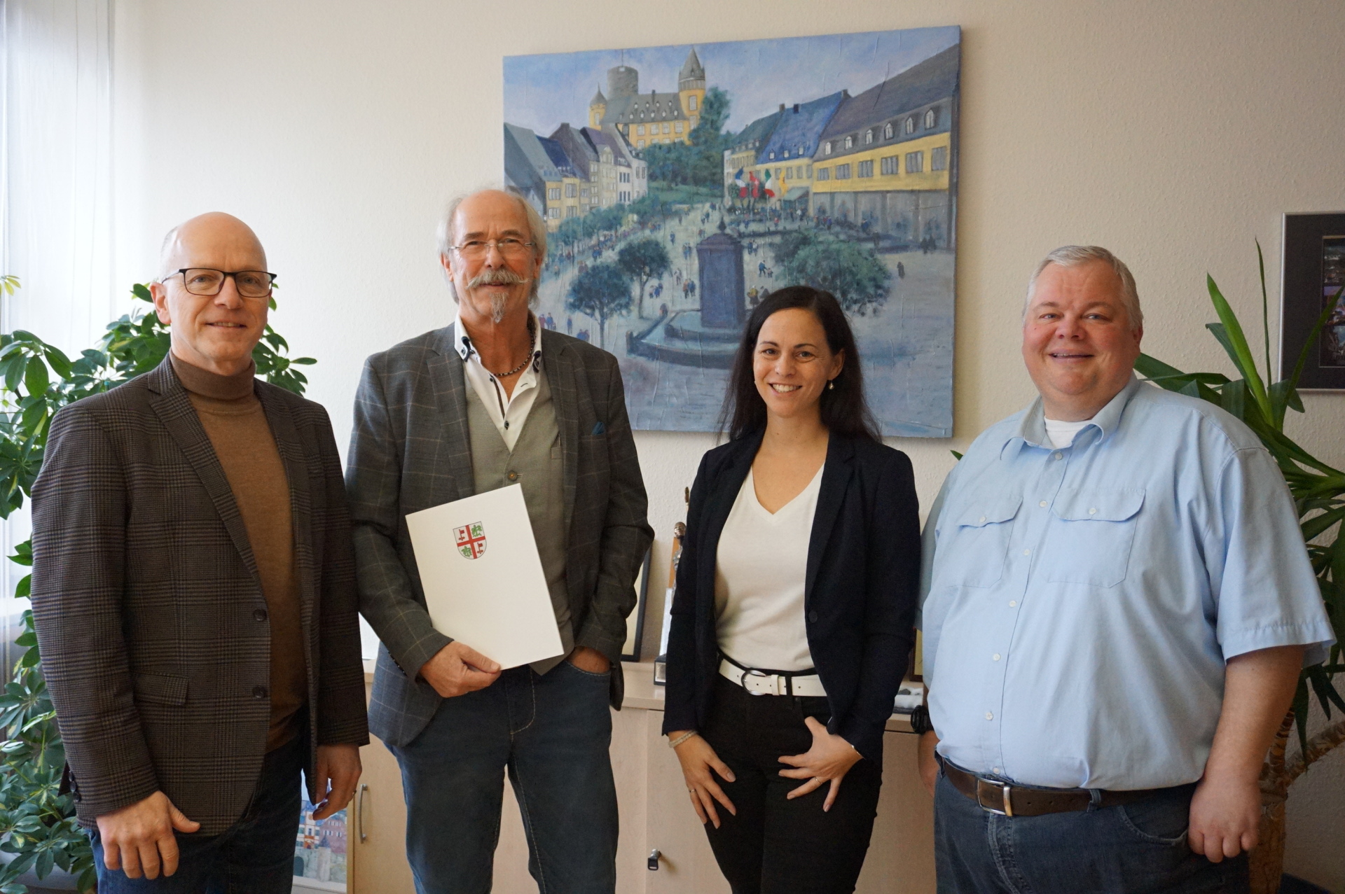 Oberbürgermeister Dirk Meid, Peter Loser, Büroleitung Jasmin Alter, Personalratsvorsitzender Wolfgang Goeken stehen gemeinsam in einem Büro. Peter Loser hält eine Urkunde in der Hand