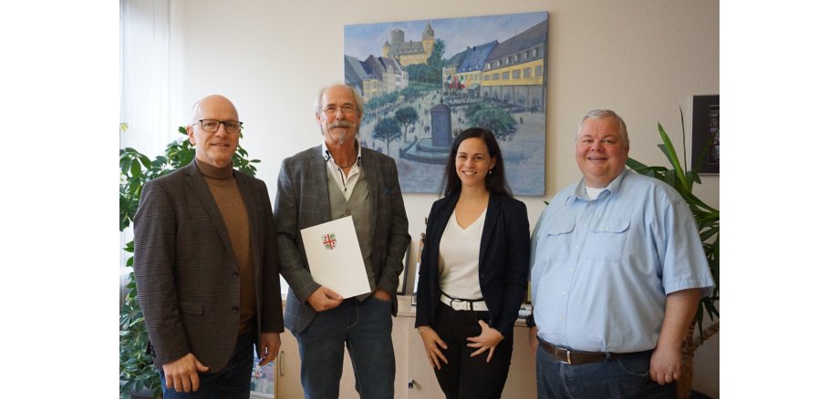 Oberbürgermeister Dirk Meid, Peter Loser, Büroleitung Jasmin Alter, Personalratsvorsitzender Wolfgang Goeken stehen gemeinsam in einem Büro. Peter Loser hält eine Urkunde in der Hand