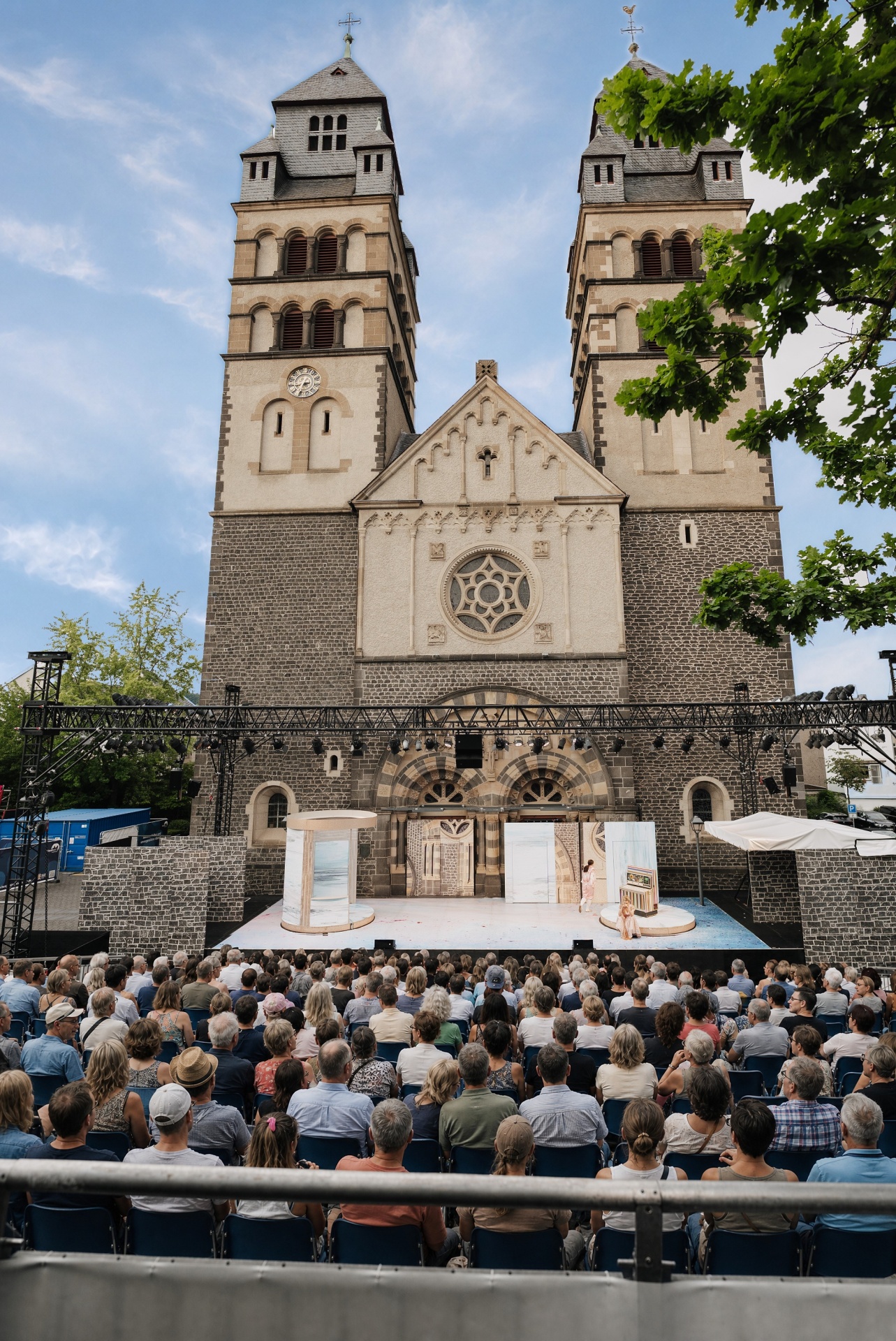 Publikum sitzt auf der Tribüne vor der Bühne der Burgfestspiele. Die Bühne steht vor der Herz Jesu Kirche.