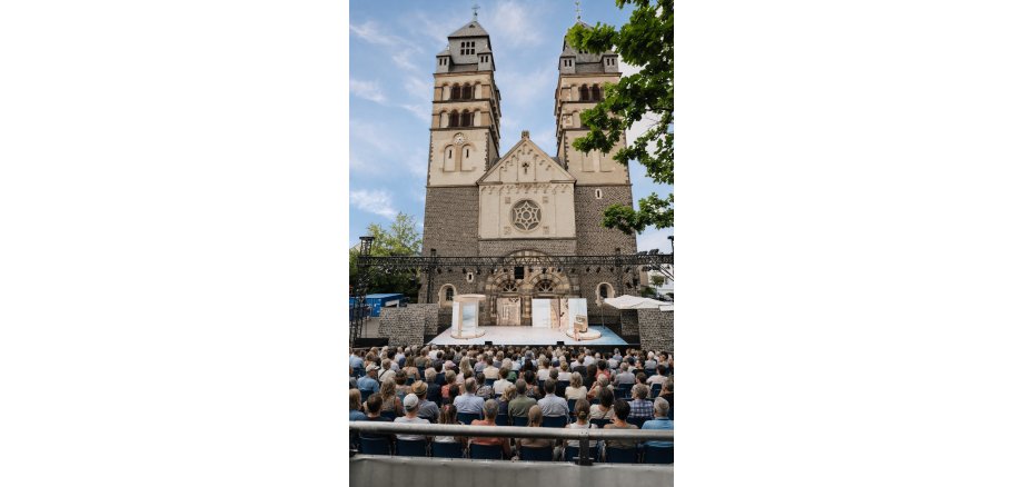 Publikum sitzt auf der Tribüne vor der Bühne der Burgfestspiele. Die Bühne steht vor der Herz Jesu Kirche.