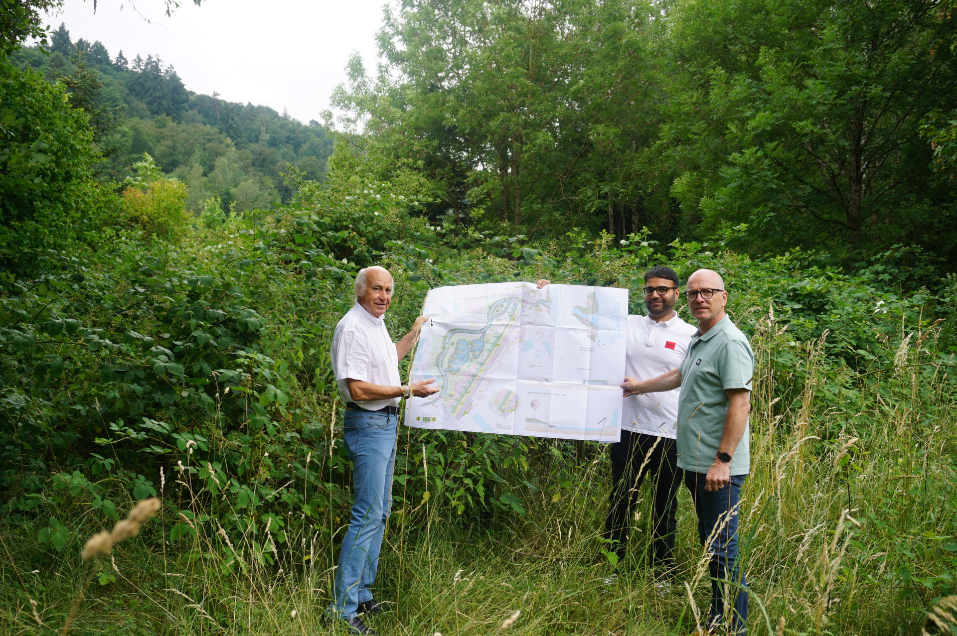 Drei Männer stehe in der Natur und halten einen Bauplan in die Luft