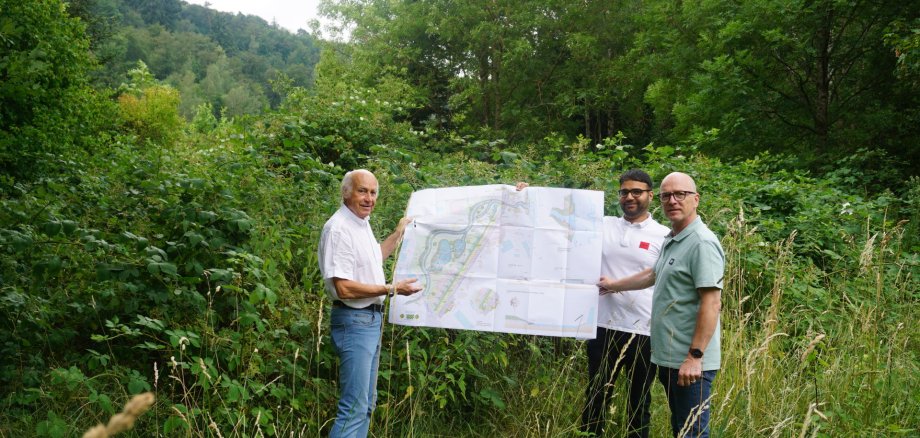Die Stadt Mayen plant die Umlegung und Renaturierung des Eiterbachs im Bereich „Stadion“. Ziel ist eine naturnahe Gewässerentwicklung und ein verbesserter Hochwasserschutz Drei Männer stehe in der Natur und halten einen Bauplan in die Luft