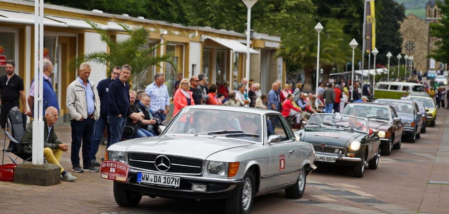 Historische Fahrzeuge der Ahr-Rotwein-Klassik machen am 28. Juni Station auf dem Mayener Marktplatz vor der Kulisse der Genovevaburg Oldtimer auf einer Straße. Am Rand stehen Zuschauer