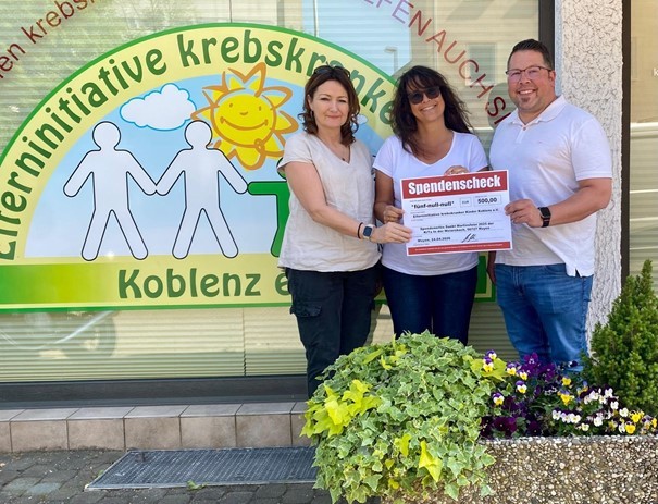 Zwei Frauen und ein Mann stehen vor einem bunten Schaufenster und halten gemeinsam einen Spendencheck in den Händen