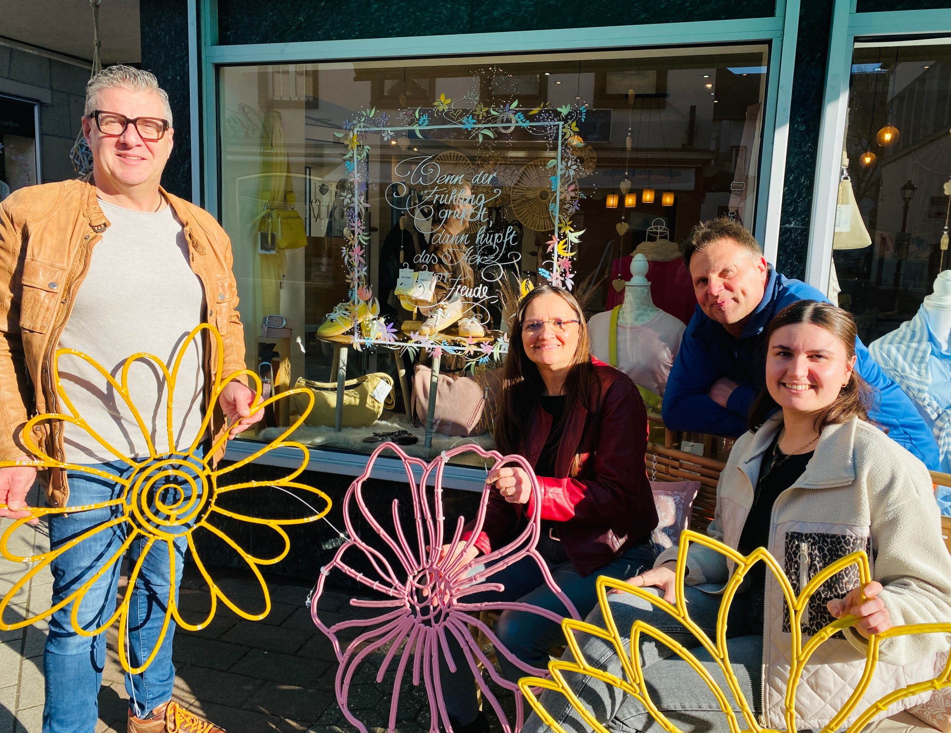 Jürgen Nett, MY-Gemeinschaft; Ayla Rozera, Brückenstraßengemeinschaft; Jörg Wagner, städtischer Betriebshof; Julia Judt, Stadt Mayen sitzen nebeneinander draußen vor einem bunten Schaufenster und halten die Blumendekoration in den Händen