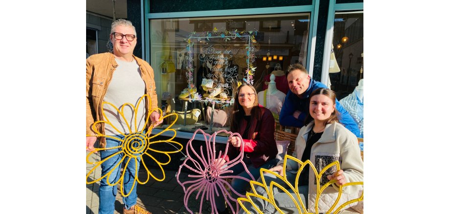 Jürgen Nett, MY-Gemeinschaft; Ayla Rozera, Brückenstraßengemeinschaft; Jörg Wagner, städtischer Betriebshof; Julia Judt, Stadt Mayen sitzen nebeneinander draußen vor einem bunten Schaufenster und halten die Blumendekoration in den Händen